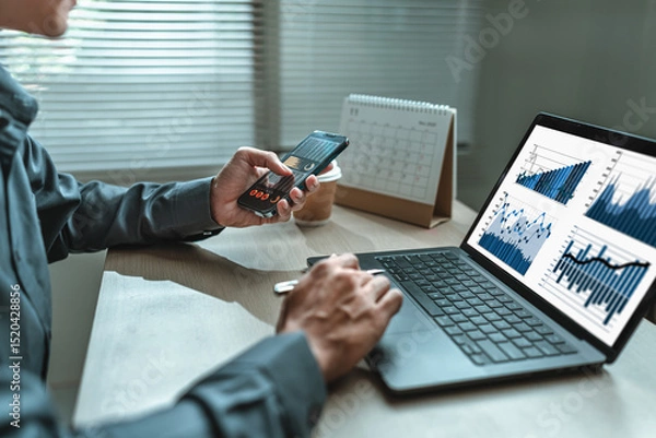Fototapeta Businessman uses mobile phone to check stocks, news, email, market information while working with laptop screen graph, chart in Office.