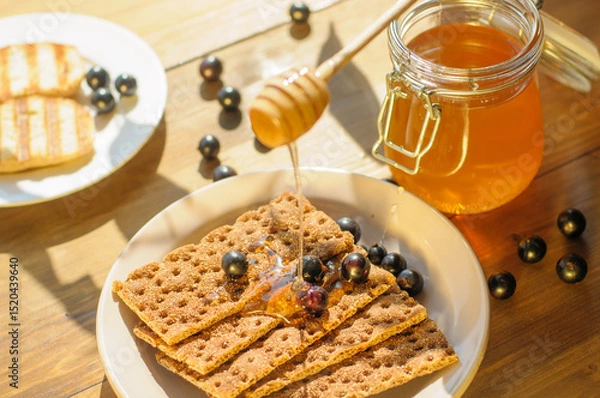 Fototapeta Golden honey drips over crispbread scattered with fresh berries, set against a rustic wooden table. The warmth of sunlight enhances the inviting atmosphere.