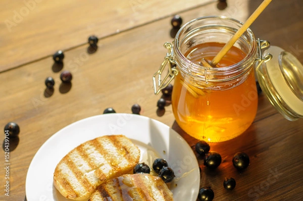Fototapeta Golden honey in a glass jar sits next to grilled bread on a white plate, with fresh berries scattered around on a rustic wooden table, evoking warmth and flavor.