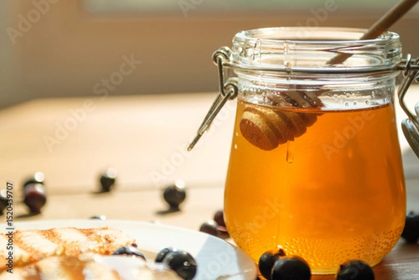 Fototapeta A wooden dipper is seen immersing in a jar of golden honey, casting delightful reflections in the sunlight, surrounded by fresh blueberries on a plate.