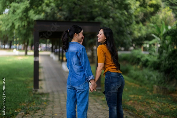 Fototapeta Two friends enjoying a leisurely stroll, holding hands and chatting happily in a green park