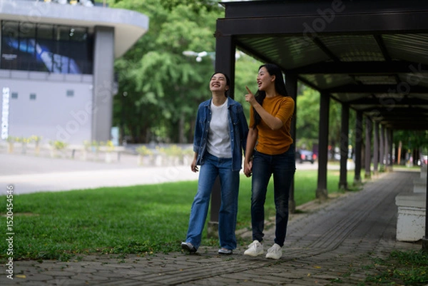 Fototapeta Two friends enjoying a leisurely stroll in a park, sharing laughter and pointing at something interesting