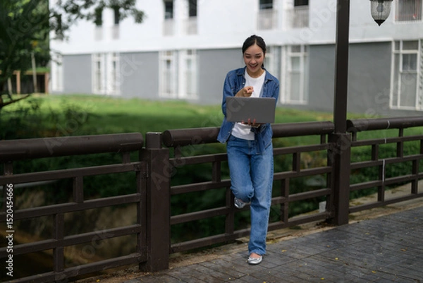 Fototapeta Young asian student pointing at laptop standing on bridge in university campus