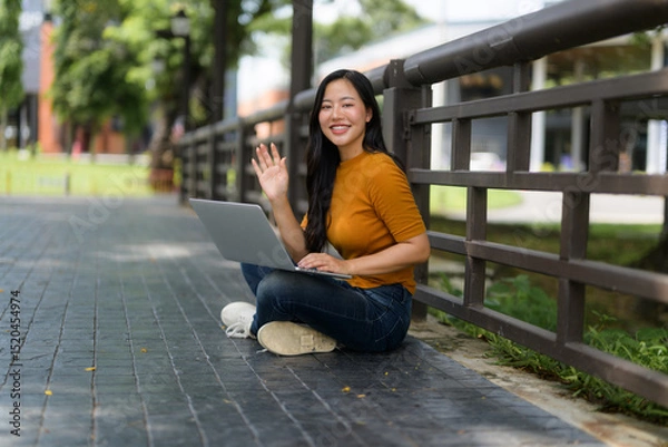 Fototapeta Freelancer working remotely having a video conference in a park, waving at the laptop screen