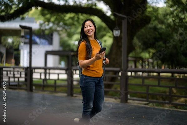 Fototapeta Asian student walking in a park using her smartphone and carrying a backpack