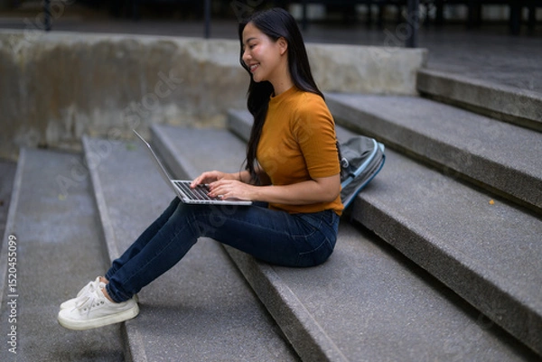 Fototapeta Smiling young Asian woman using laptop sitting on campus stairs, enjoying online learning and studying outdoors