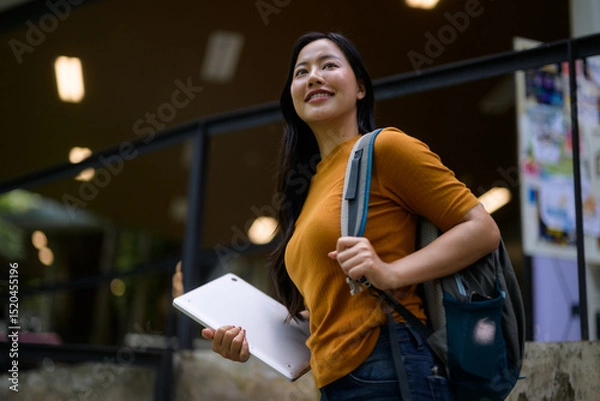 Fototapeta Young Asian student walking up stairs holding tablet and carrying backpack at university