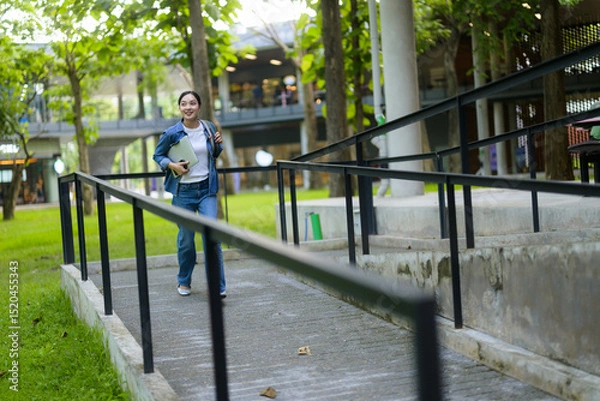 Fototapeta Asian female college student going up an accessible ramp in a university campus, holding a laptop, and smiling