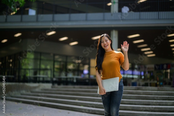 Fototapeta Happy asian student waving while holding laptop in university campus