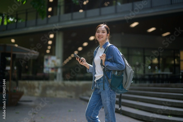 Fototapeta Asian female student using mobile phone and carrying backpack walking on university campus