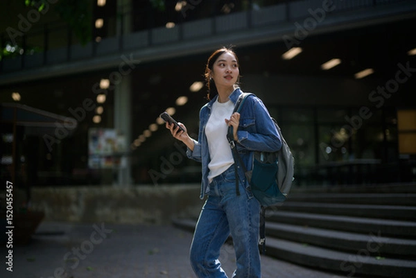 Fototapeta University student holding her smartphone while walking outside university campus building