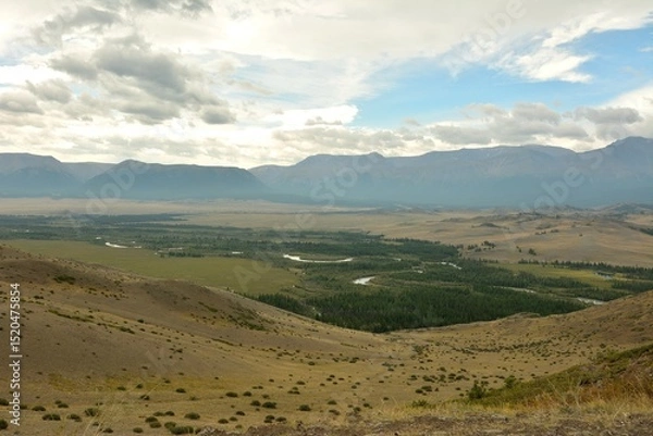 Obraz A wide, winding river crosses a vast steppe lying at the foot of a snow-capped mountain range on a cloudy summer day.