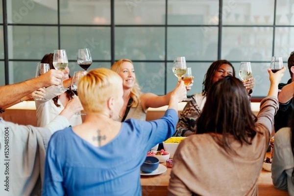 Fototapeta Group of diverse people raising glasses in celebration. Mixed gender and ethnicity. Smiling faces, joyful atmosphere, and toasting in a lively gathering. People in festive dinner, celebration.