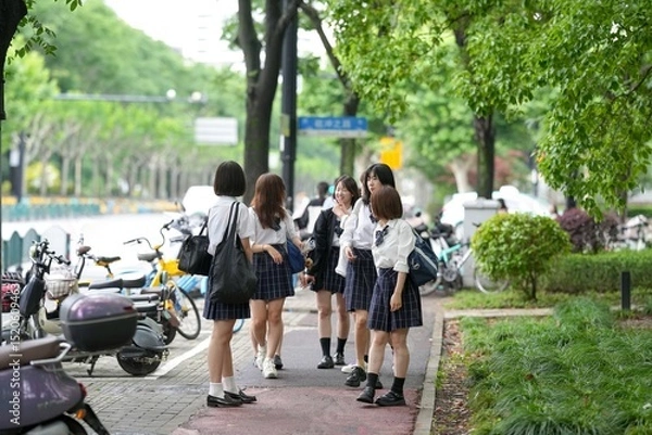 Fototapeta In early June in Pudong, Shanghai, seven Japanese high school girls in summer uniforms walk together down a city street, smiling and chatting as they head to school under the soft morning sunlight.