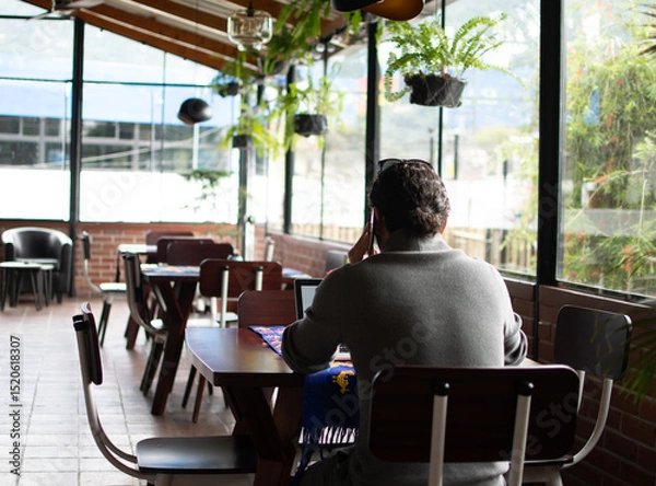 Fototapeta Photograph of an adult man talking on the phone in a cafe in front of a laptop at a wooden table with a textile tablecloth, with other tables covered in fabric, seen from behind.