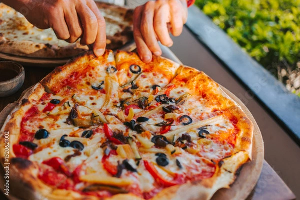 Fototapeta Men's hands tear off a slice of pizza with vegetable toppings, olives and stretching hot stretchy cheese. Process of eating pizza close-up. Delicious Italian pizza for friends and delivery