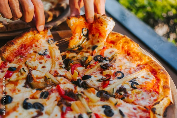 Fototapeta Men's hands tear off a slice of pizza with vegetable toppings, olives and stretching hot stretchy cheese. Process of eating pizza close-up. Delicious Italian pizza for friends and delivery