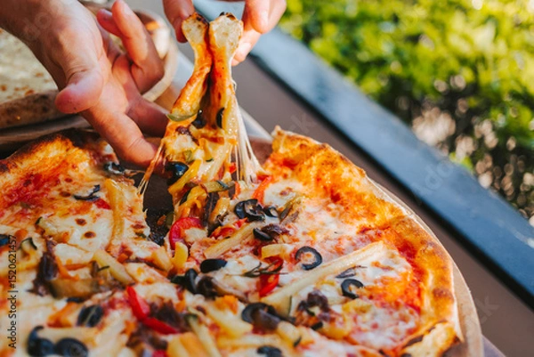 Fototapeta Men's hands tear off a slice of pizza with vegetable toppings, olives and stretching hot stretchy cheese. Process of eating pizza close-up. Delicious Italian pizza for friends and delivery