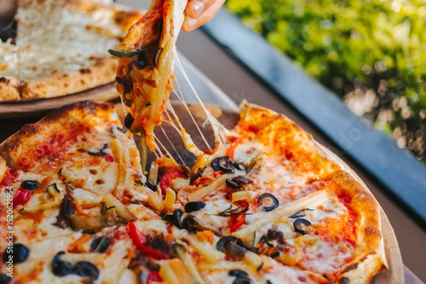 Fototapeta Men's hands tear off a slice of pizza with vegetable toppings, olives and stretching hot stretchy cheese. Process of eating pizza close-up. Delicious Italian pizza for friends and delivery