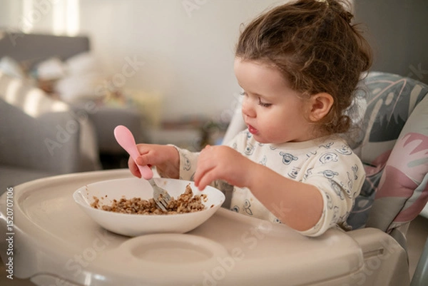 Fototapeta Happy toddler with curls having breakfast in a high chair. Kitchen background with bowl, spoon, and loving family environment.