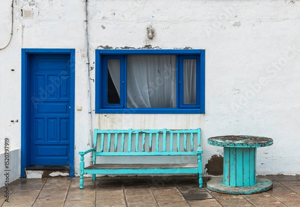 Fototapeta Weathered wooden bench with faded turquoise paint sits on a paved patio. Adjacent is a vivid blue door set in a white wall. A matching blue-framed window with net curtains is above the bench.