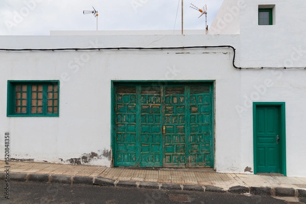 Fototapeta White building with an old, weathered green wooden door featuring a detailed geometric pattern. Accompanied by a matching smaller green door on the right and a window with green shutters on the left.