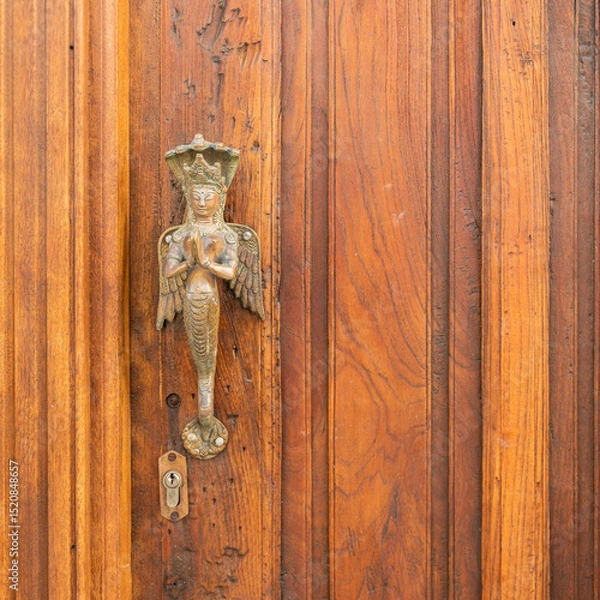 Fototapeta Ornate brass door handle features a figure resembling a celestial being or deity, with folded hands and decorative attire, possibly inspired by Southeast Asian art. The figure has wings.