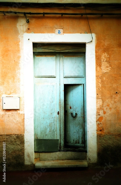 Fototapeta A weathered, light blue wooden door with two panels in a worn, peach-colored wall. The plaster is aged, showing patches and discoloration. A white stone frame surrounds the door.