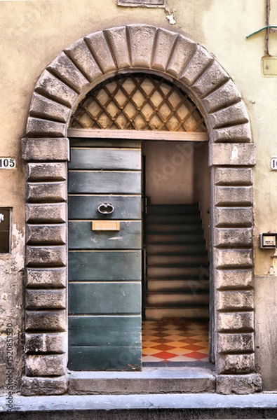 Fototapeta Arched stone doorway with a worn texture frames a partially open green wooden door. The door has simple metal hardware and a mail slot. Inside, a staircase with ornate red and yellow checkered tiles.