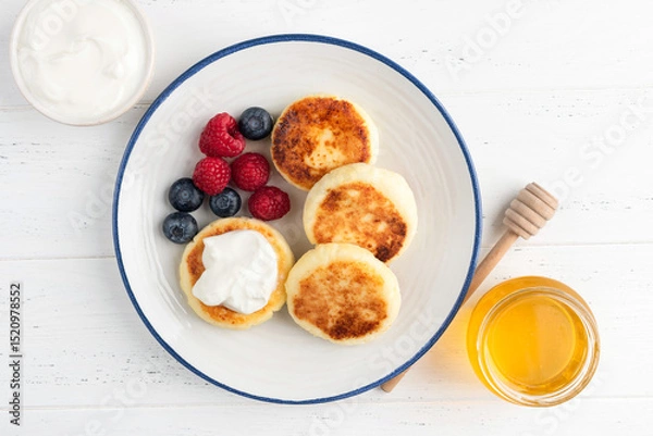 Fototapeta Cottage cheese fritters, syrniki served with fresh berries blueberry raspberry, honey and sour cream on white plate, top view