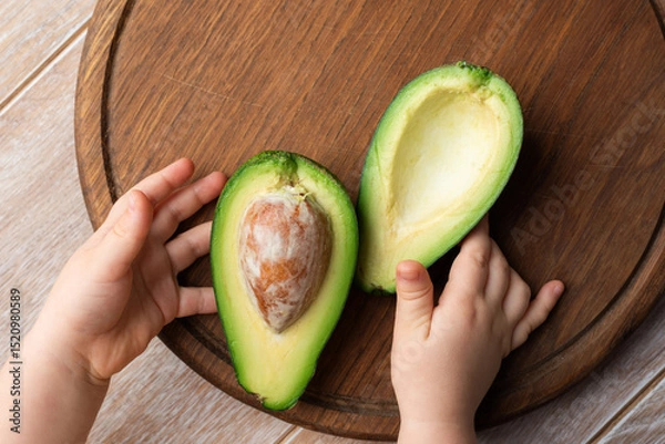 Fototapeta Avocado halves on a wooden cutting board and toddler hands, top view