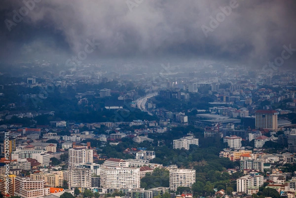 Obraz aerial view of chiang mai cityscape under a cloudy, rainy season sky, with urban buildings, a winding road, and green trees partially obscured by mist.
