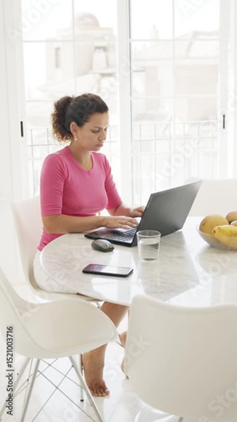 Fototapeta Middle aged woman is sitting at the table and working on a laptop at home.  White colored interior of apartment.