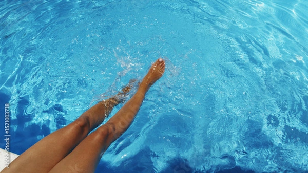 Fototapeta Woman is sitting at the edge of the pool with her legs dangling in the water and shaking the water  and making ripples and small waves. Summer background.