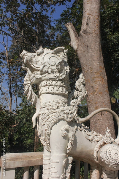 Fototapeta Decorative White Singha at Temple Entrance. Side view of a white Singha guardian statue with elaborate carvings, located at the gate of a Thai temple under bright sunlight.