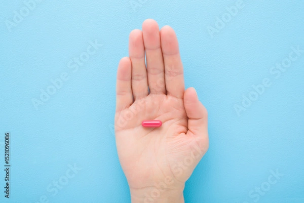 Fototapeta Young adult woman opened palm holding and showing red pill for improve of iron level in blood on light blue table background. Pastel color. Closeup. Top down view.