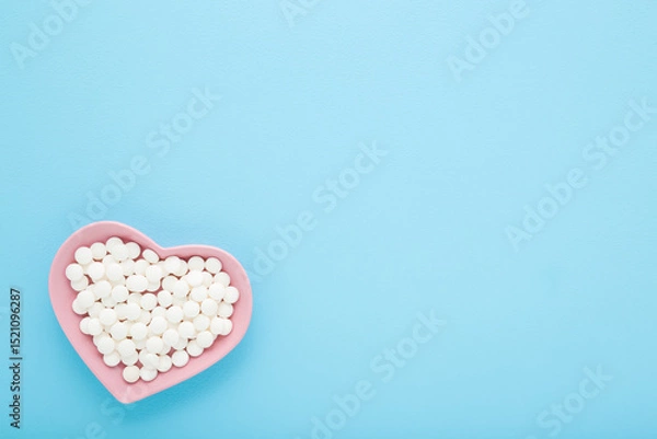 Fototapeta White pills in pink heart shape plate on light blue table background. Pastel color. Closeup. Healthcare concept. Empty place for text. Top down view.