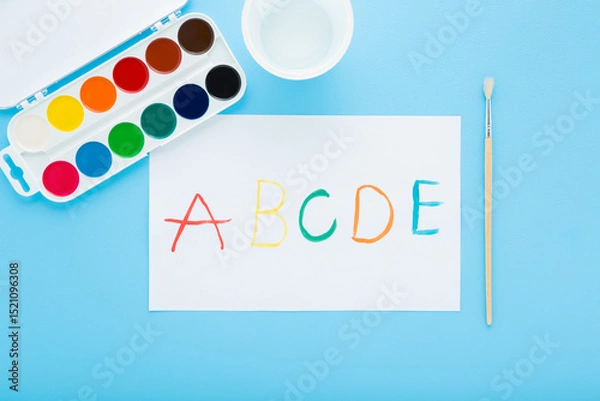 Fototapeta Colorful watercolor palette, paintbrush and drawn different english letters on white paper on light blue table background. Pastel color. Closeup. Creativity learning for little kids. Top down view.