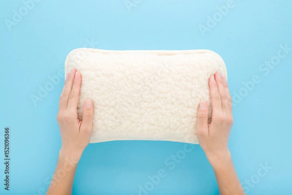 Fototapeta Young adult woman hands touching new clean white soft natural sheep wool pillow on light blue table background. Pastel color. Closeup. Point of view shot. Top down view.