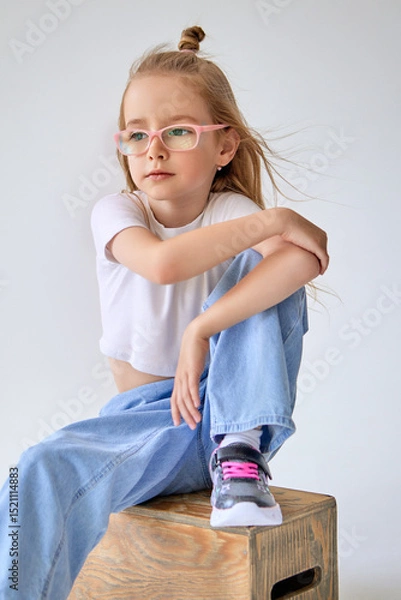 Fototapeta A young girl with glasses and long hair blowing in the wind, tied into a small bun on top, sits on a wooden box with a thoughtful expression on her face. The child sits with his hands folded on knees.