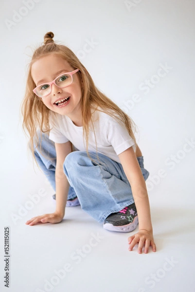 Fototapeta Mischievous young girl with long hair tied in a bun on top of her head sits on her haunches. Cheerful child in glasses laughs and touching the floor with her hands on a minimalist white background