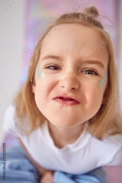 Fototapeta A little cute girl is making a funny face. The child has a mischievous expression, her eyes are squinted and her lips are slightly pouted, creating a playful look. There are glitters on her cheeks