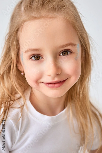 Fototapeta Close-up portrait of a lovely little girl with long blond hair, wearing a white T-shirt. Blue glitter sparkles on her cheeks. The child smiles, creating a friendly and joyful impression.