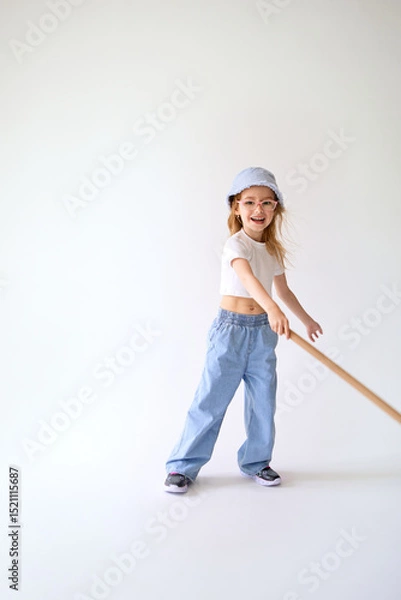 Fototapeta Young active girl in white crop top and wide jeans vigorously swings bat. She laughs and holds long stick in hands, stands in a dynamic pose on light background