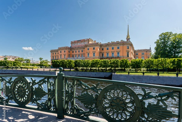Fototapeta Mikhailovsky Castle is visible behind an openwork bridge fence and a river under a blue sky.