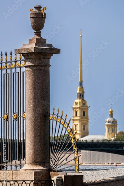 Fototapeta A close-up of a granite column and a forged grille of the Summer Garden fence, with the spire of the Peter and Paul Fortress visible in the background.