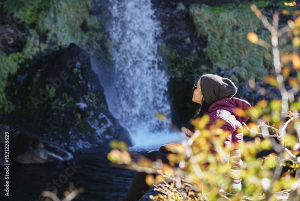Fototapeta Woman in a beanie and winter coat sits contemplatively on rocks near the base of a waterfall, surrounded by autumn foliage