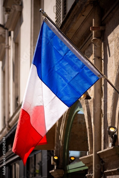 Fototapeta French flag. National colors: blue, white and red. Hanging on a flag pole in Paris (France). Tricolore “bleu blanc rouge“ blown by wind with blurred facade of an old houses and backlit by sunlight.