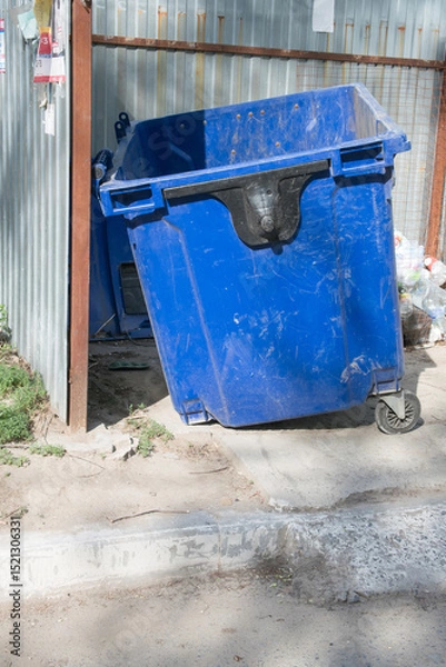 Fototapeta Large blue dumpster positioned at an urban alleyway, surrounded by a metal fence and concrete pavement, showcasing waste management and environmental concerns in city life