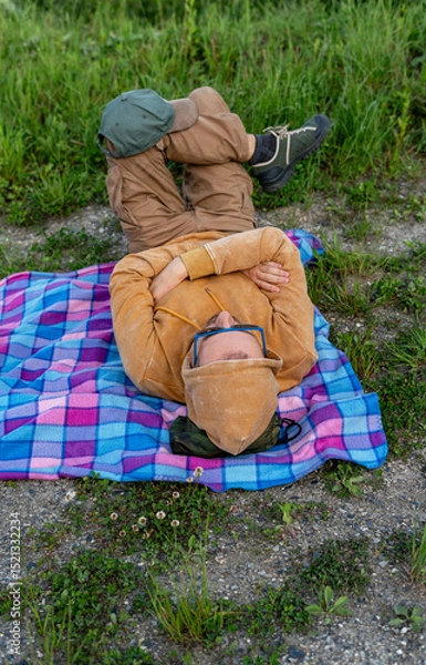 Fototapeta Top view of a man resting in yellow hoodie on a colorful checkered blanket on the ground, surrounded by grass and nature, hiking and nature exploration